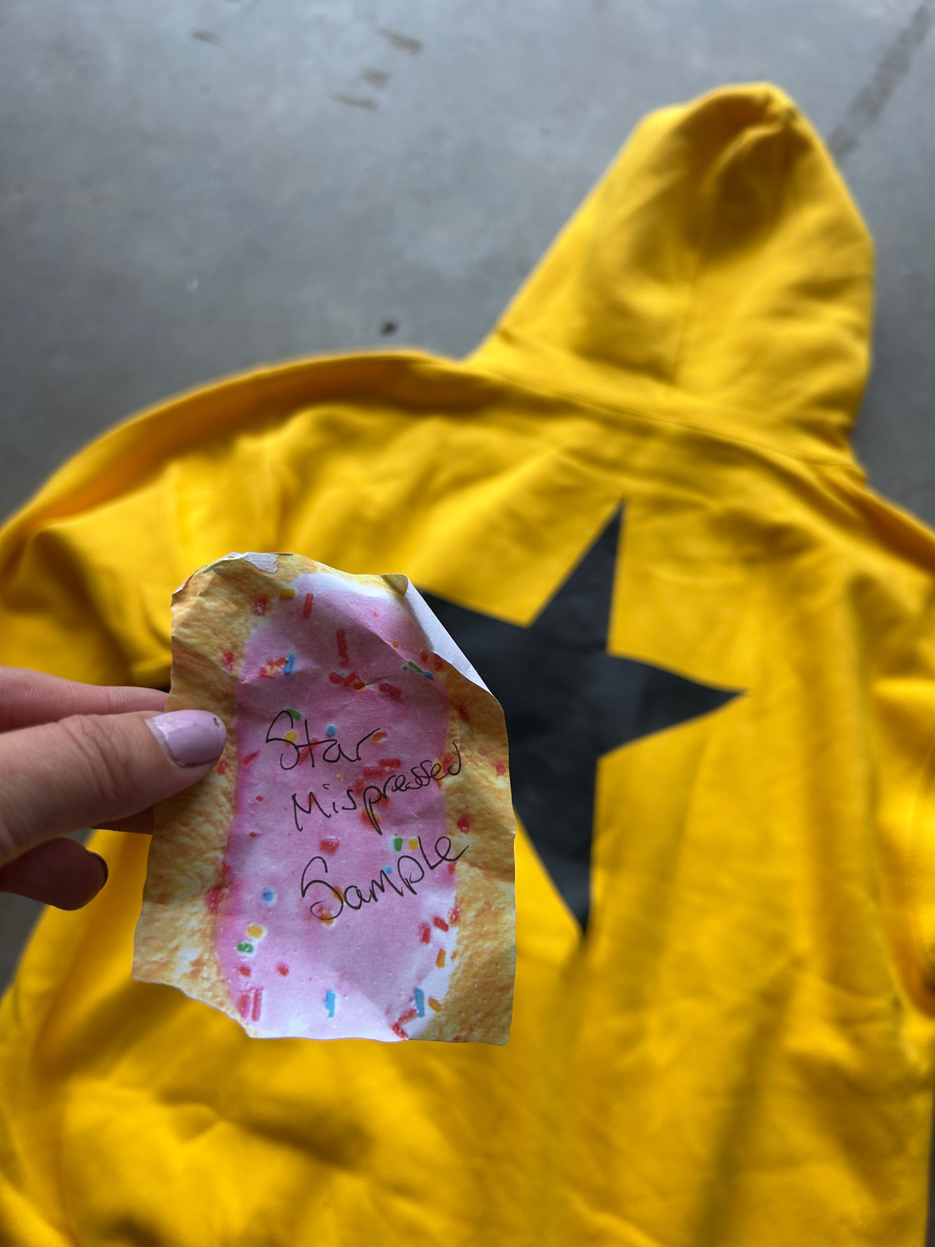 A hand holds a small pink and yellow novelty tag that reads 'Star Misplaced Sample' in front of a bright yellow hoodie with a large black star on the back, set on a gray concrete floor.