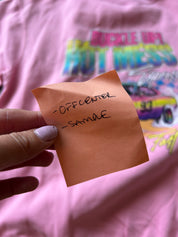 Hand holding a sticky note that reads "off center' and 'sample; with a pink crewneck in the background