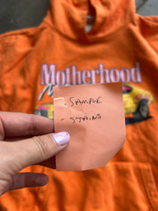 A hand holds a small peach-colored sticky note in front of an orange hoodie with a 'Motherhood' graphic. The sticky note is handwritten with the words 'SAMPLE' and 'STAINS'.