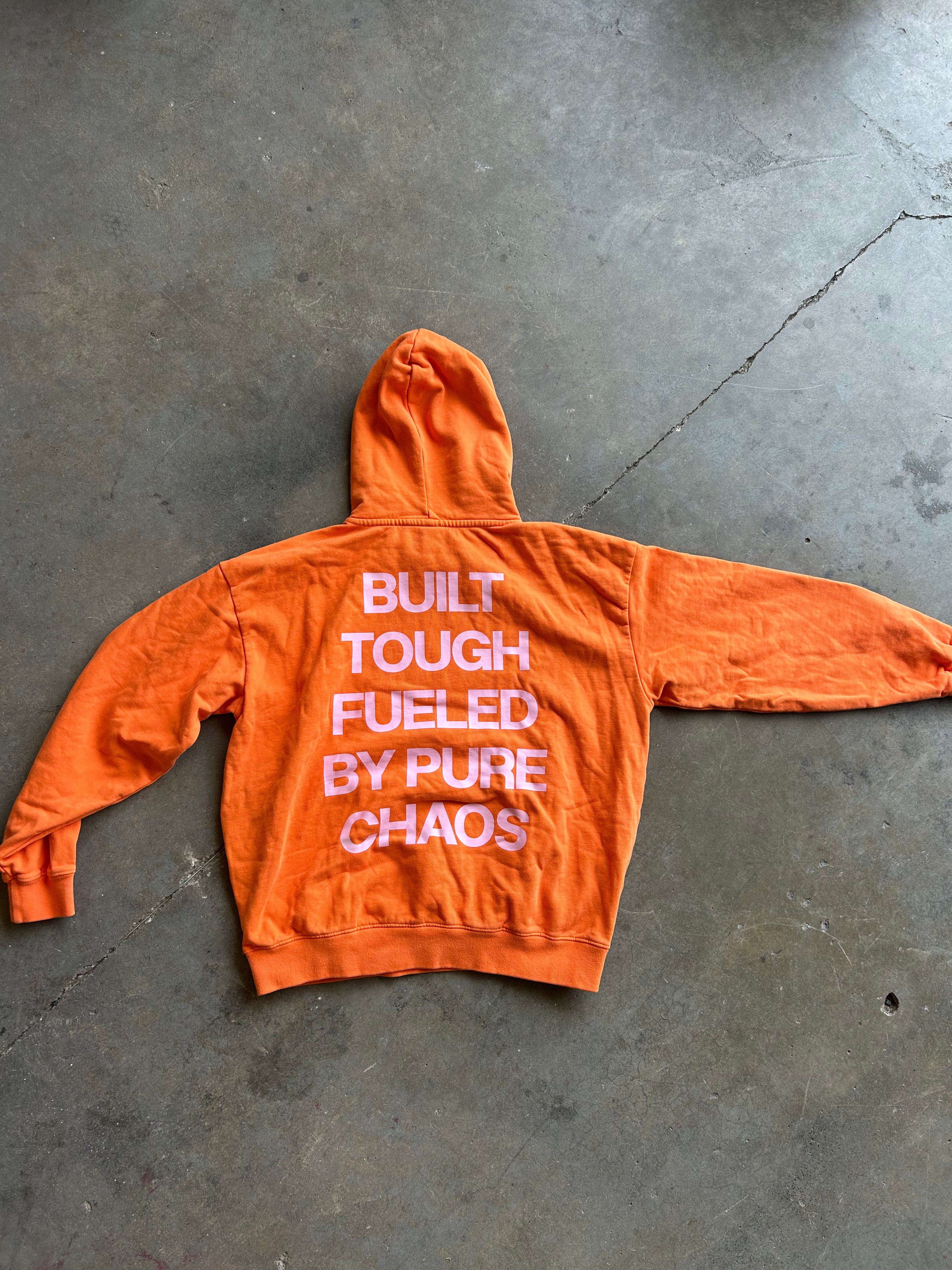 An orange hooded sweatshirt laid flat on a gray concrete floor, showing the back with large white text that reads 'BUILT TOUGH FUELED BY PURE CHAOS'.