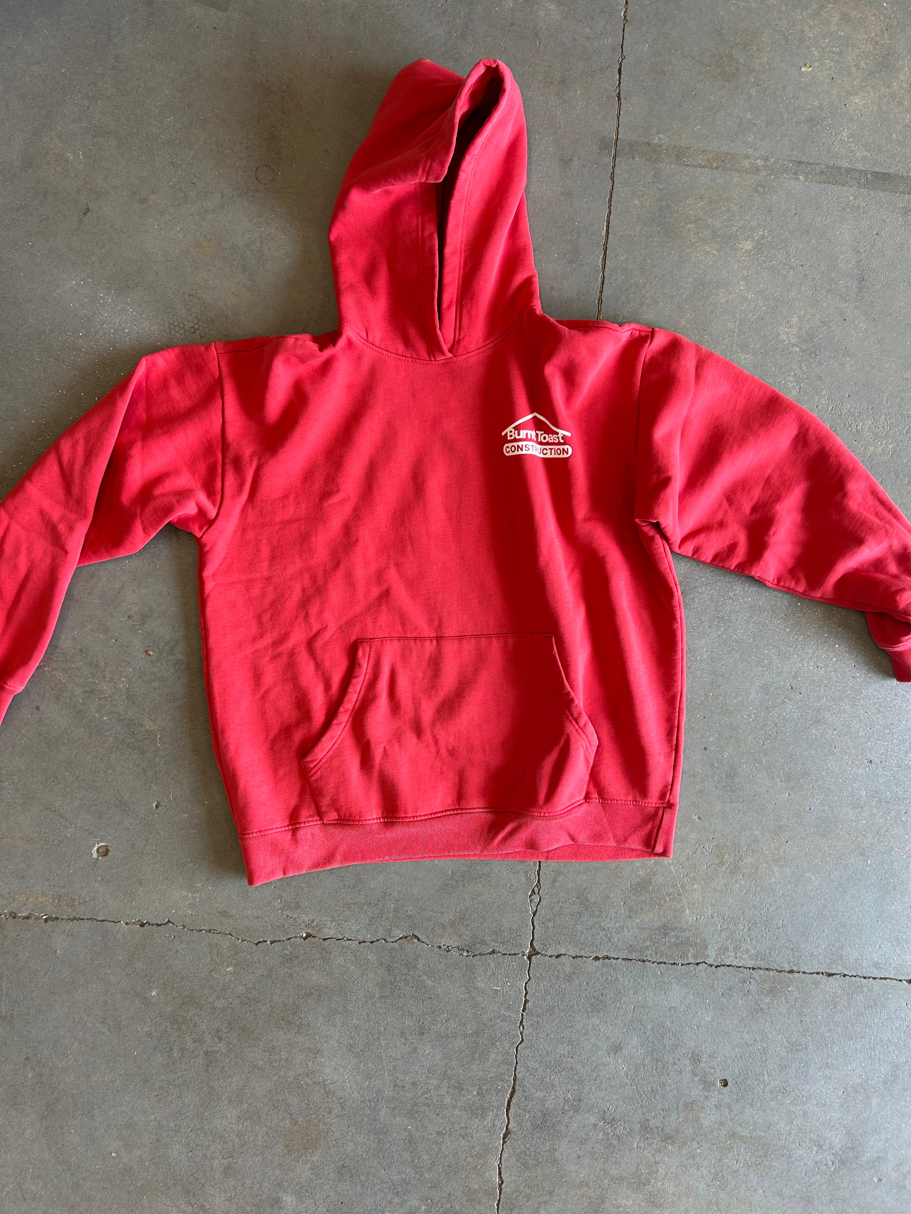 A red pullover hoodie with a small white graphic logo on the left chest, laid flat on a gray concrete floor.