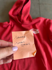 A hand with lavender nail polish holds a small orange sticky note that reads 'SAMPLE - SMALL STAINS' over a red hooded sweatshirt.