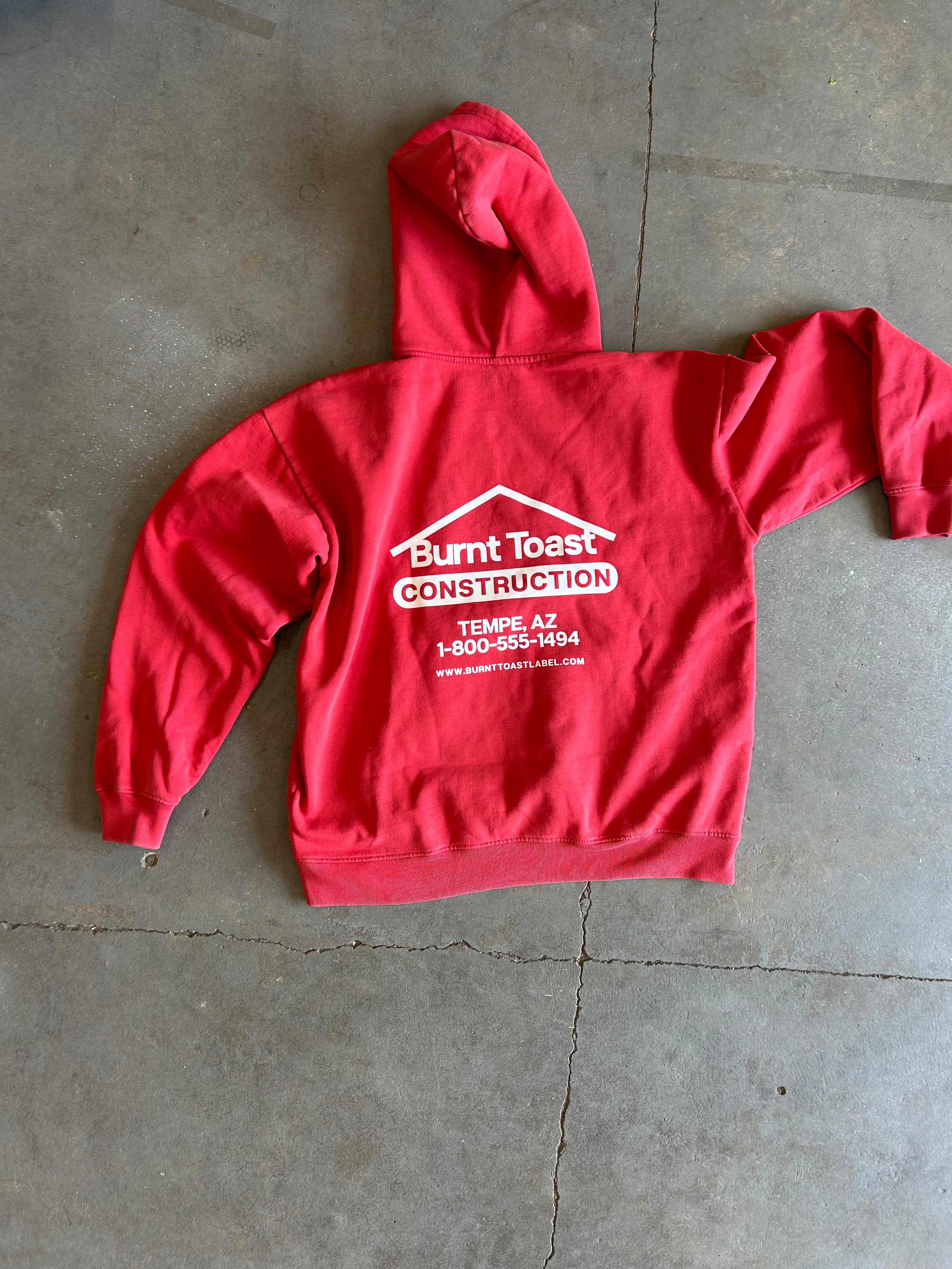 A red pullover hoodie laid flat on a gray concrete floor, showing the back side with a white printed logo that reads 'Burnt Toast Construction' and a phone number.