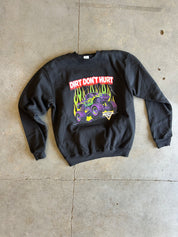 A black crewneck sweatshirt laid flat on a concrete floor, featuring a colorful graphic of a purple monster truck with yellow flames and the text 'DIRT DON'T HURT' in red and white.