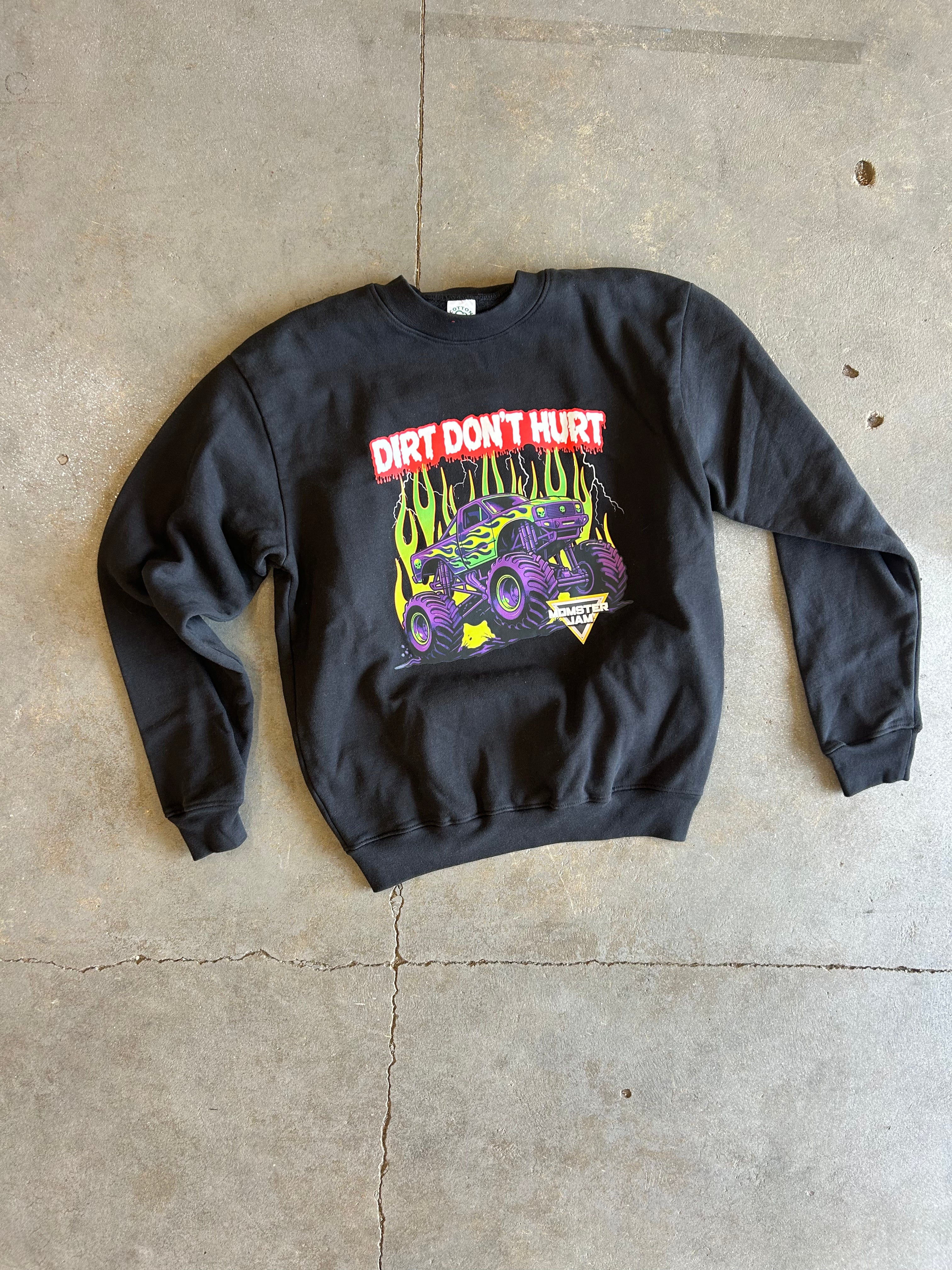 A black crewneck sweatshirt laid flat on a concrete floor, featuring a colorful graphic of a purple monster truck with yellow flames and the text 'DIRT DON'T HURT' in red and white.