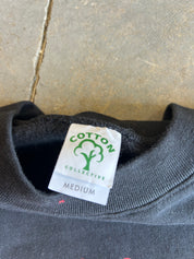 Close-up of a dark gray crewneck sweatshirt showing the inner neck label which reads 'COTTON COLLECTIVE' with a green cotton boll logo and 'MEDIUM' size text, set against a gray tiled background.