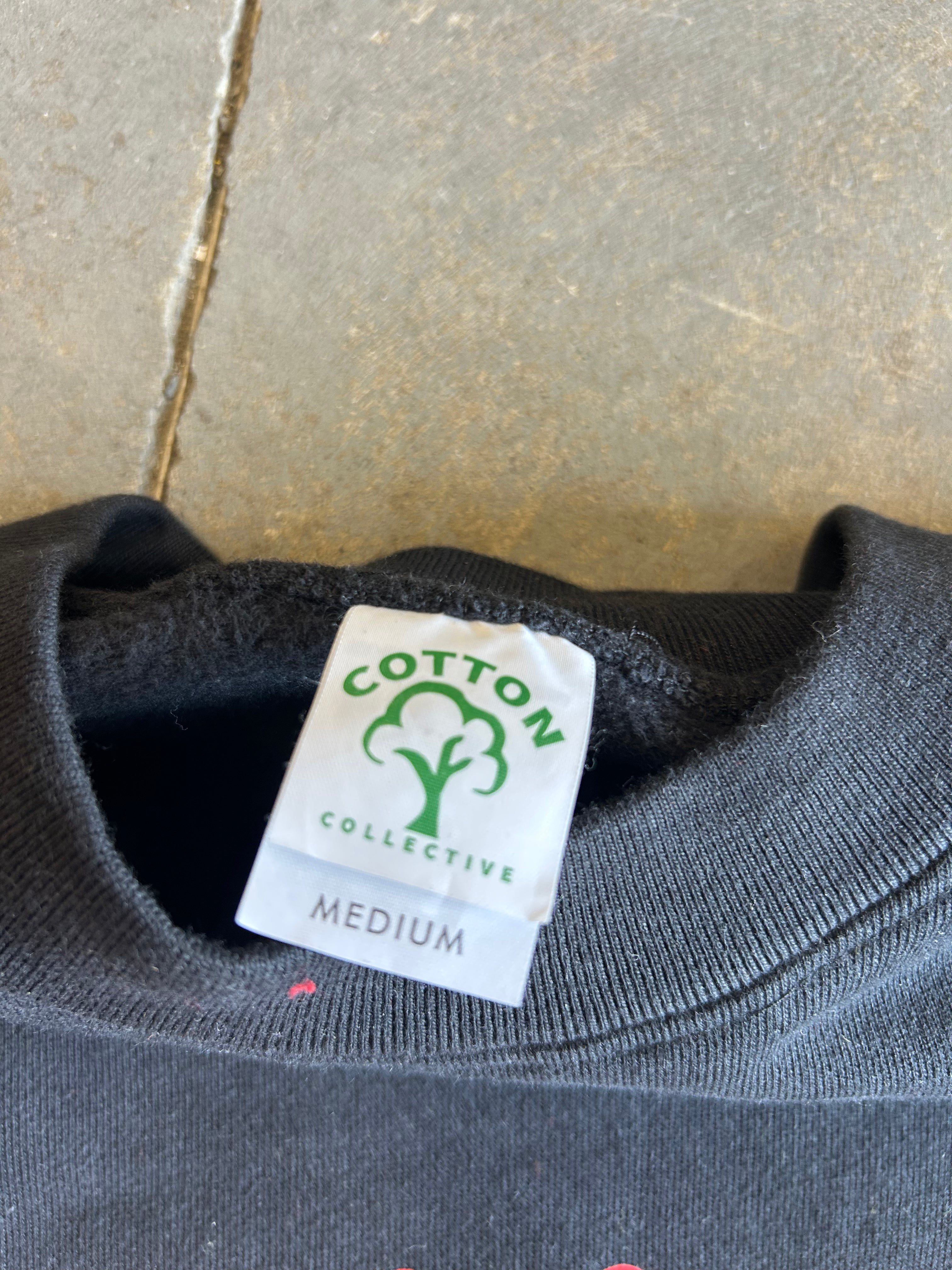 Close-up of a dark gray crewneck sweatshirt showing the inner neck label which reads 'COTTON COLLECTIVE' with a green cotton boll logo and 'MEDIUM' size text, set against a gray tiled background.