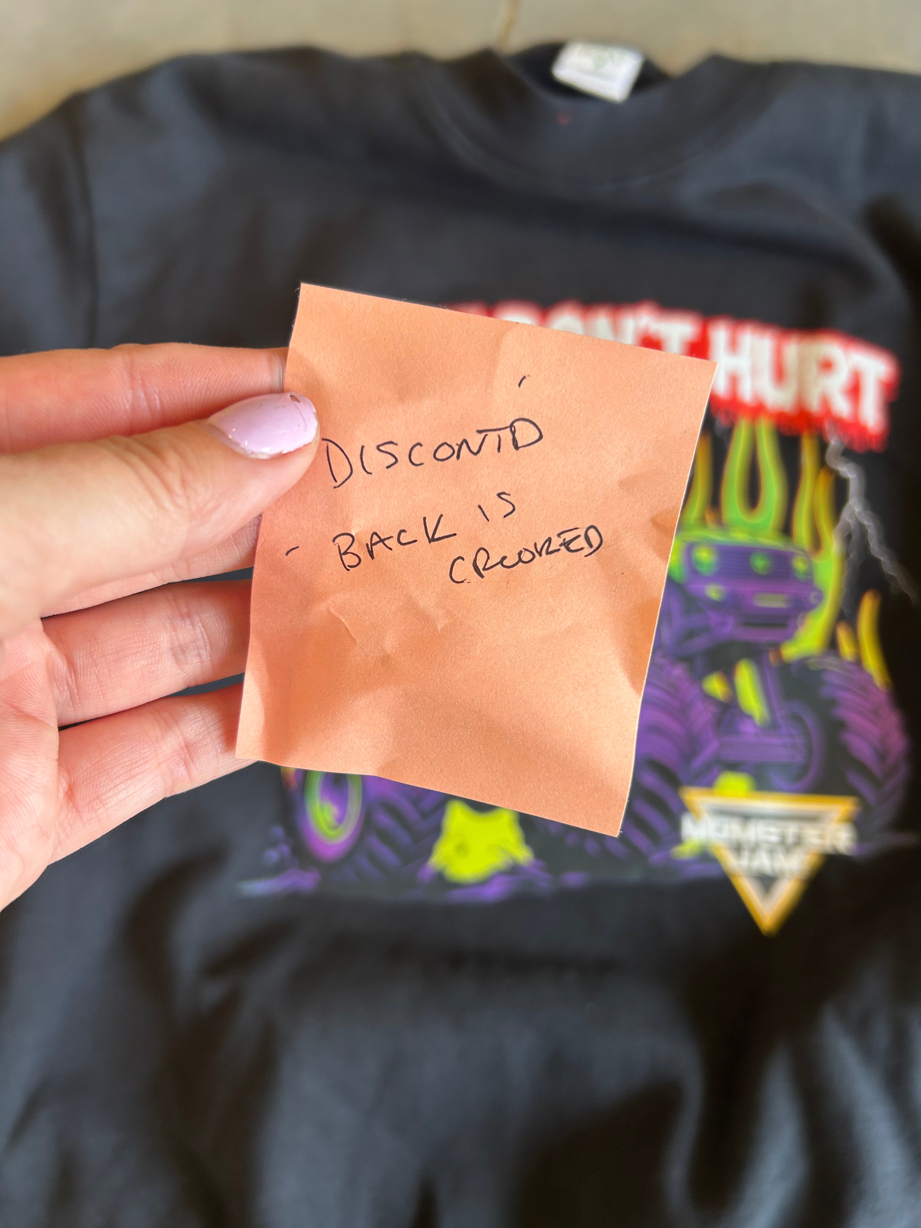 A hand with lavender nail polish holds a handwritten orange sticky note in front of a black graphic t-shirt. The note reads 'DISCOUNT - BACK IS CROOKED'.