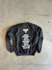 A black crewneck sweatshirt laid flat on a concrete floor, showing the back side with white text listing various mother figures and a small yellow and white graphic at the top.