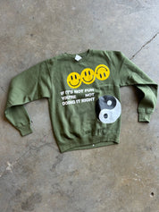 An olive green crewneck sweatshirt laid flat on a concrete floor. The sweatshirt features three yellow smiley faces, the text 'IF IT'S NOT FUN YOU'RE NOT DOING IT RIGHT' in white, and a black and white yin-yang symbol.