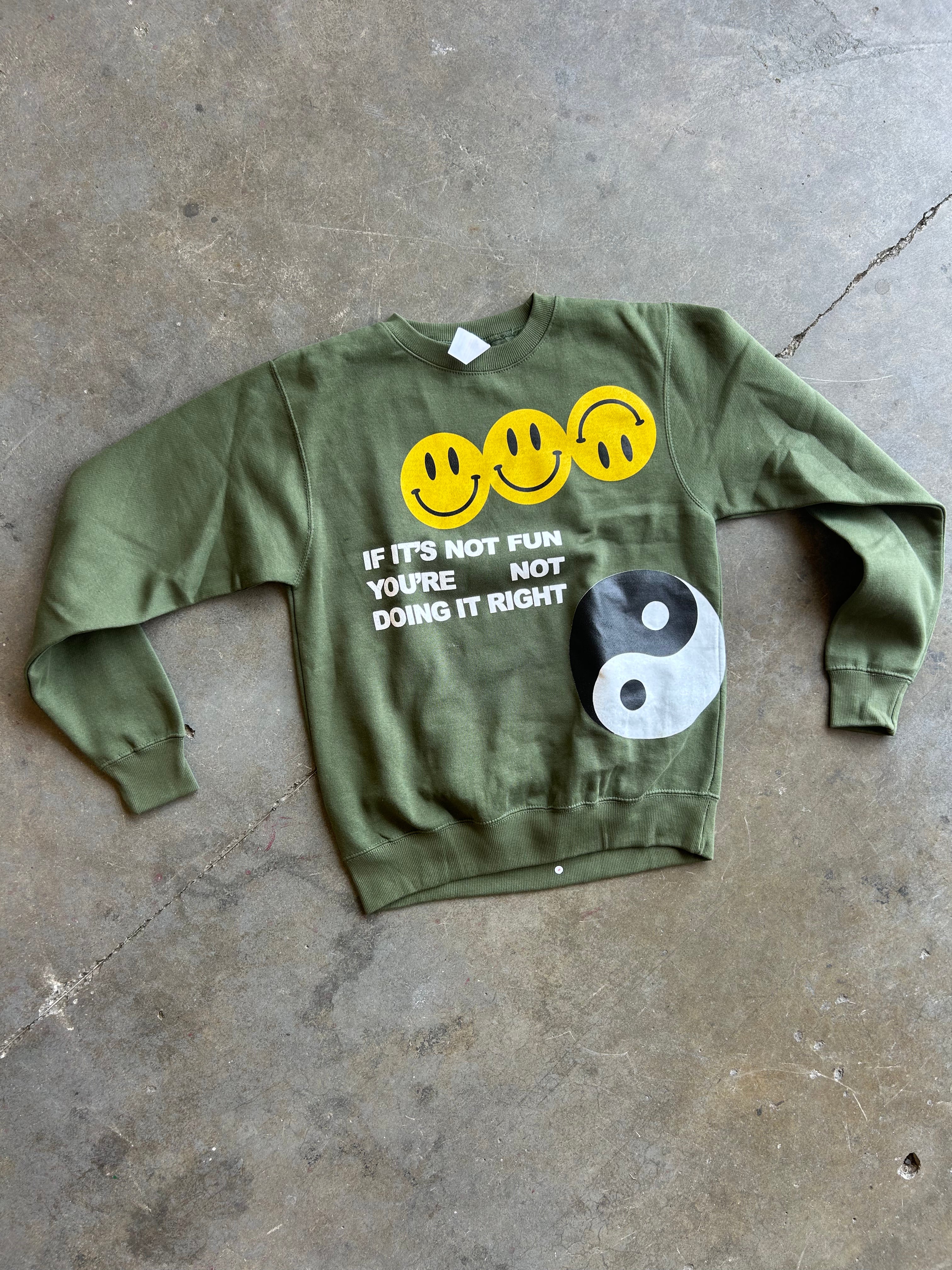 An olive green crewneck sweatshirt laid flat on a concrete floor. The sweatshirt features three yellow smiley faces, the text 'IF IT'S NOT FUN YOU'RE NOT DOING IT RIGHT' in white, and a black and white yin-yang symbol.