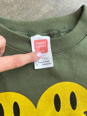 Close-up of an olive green sweatshirt with a yellow smiley face graphic at the bottom and a finger pointing to a white tag that reads 'THREE LAYER' and 'Sweatshirt'.