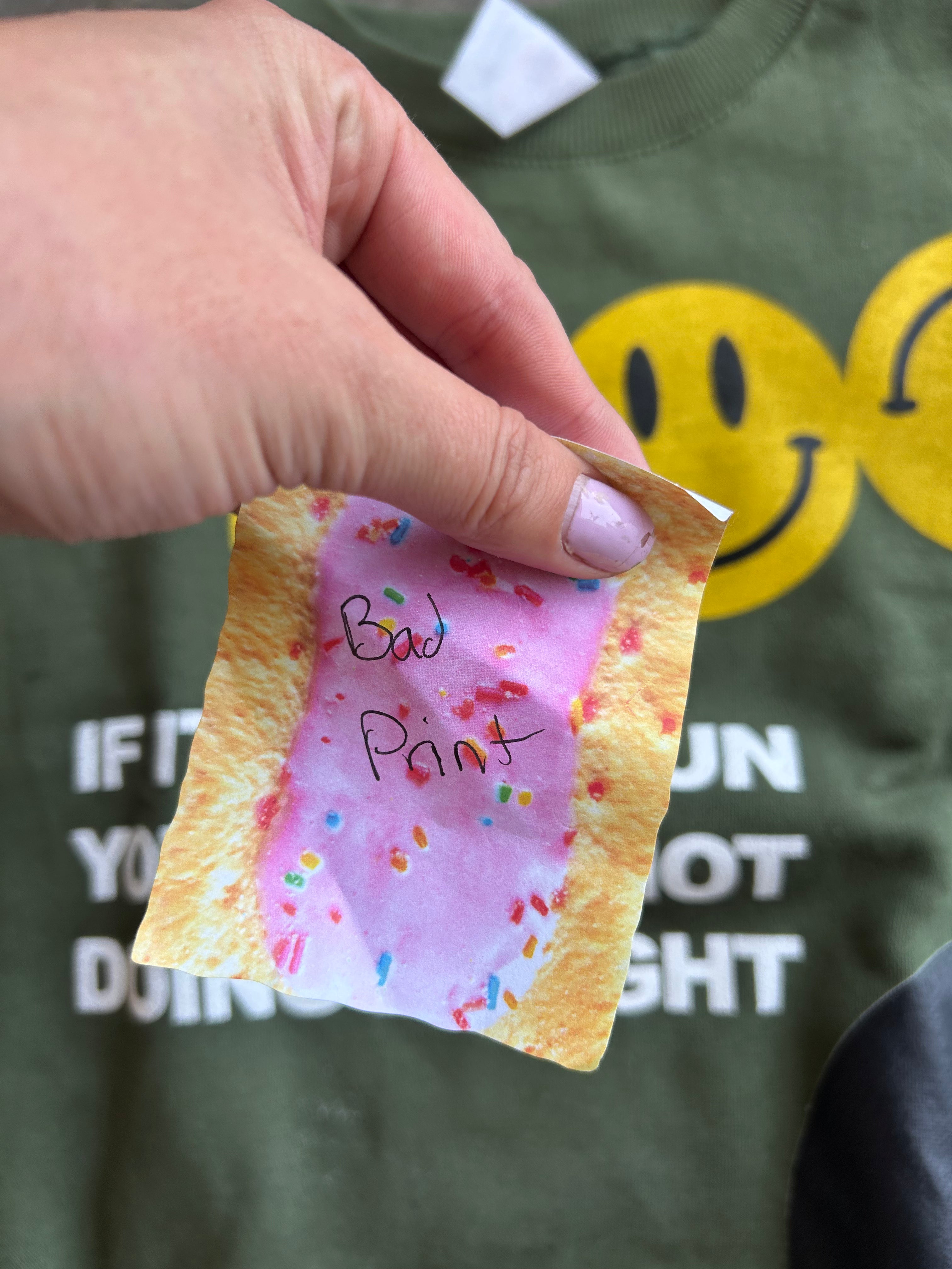 A hand holding a small printed card or sticker shaped like a pink frosted pastry with sprinkles and handwritten text reading 'Bad Print' in front of an olive green t-shirt with yellow smiley faces and white text.