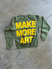 An olive green crewneck sweatshirt laid flat on a concrete floor, featuring the phrase 'MAKE MORE ART' printed in bold yellow letters across the back.