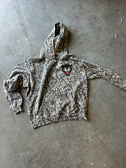 A camouflage hoodie with a small embroidered panther logo on the chest, laid flat on a textured gray concrete floor.