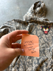 A hand holds an orange sticky note in front of a gray and black snakeskin-patterned hoodie. The hoodie has a small embroidered panther face patch on the chest.