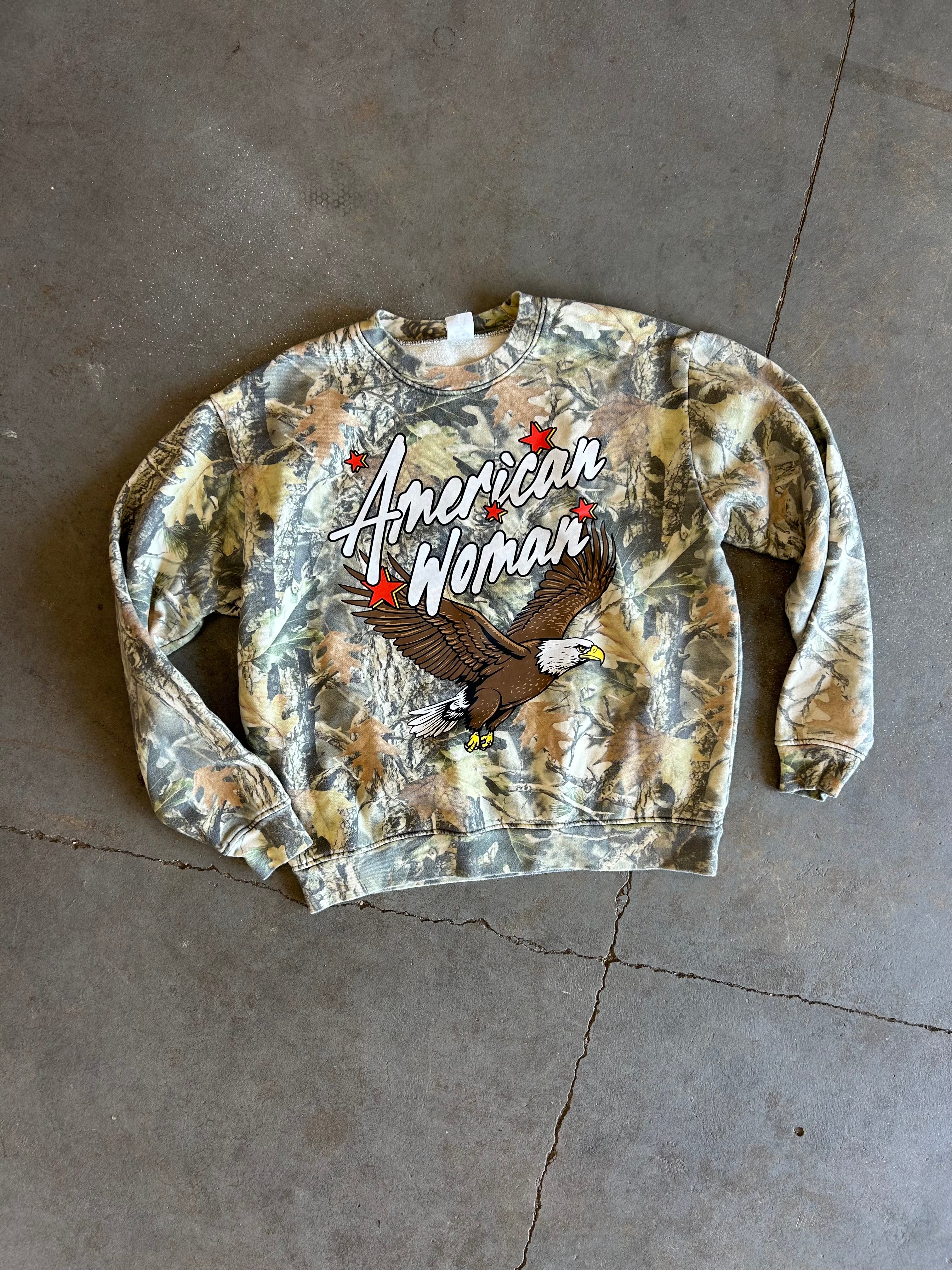 A long-sleeve crewneck sweatshirt with a camouflage pattern, featuring a graphic of a bald eagle and the text 'American Woman' with red stars, laid flat on a concrete floor.
