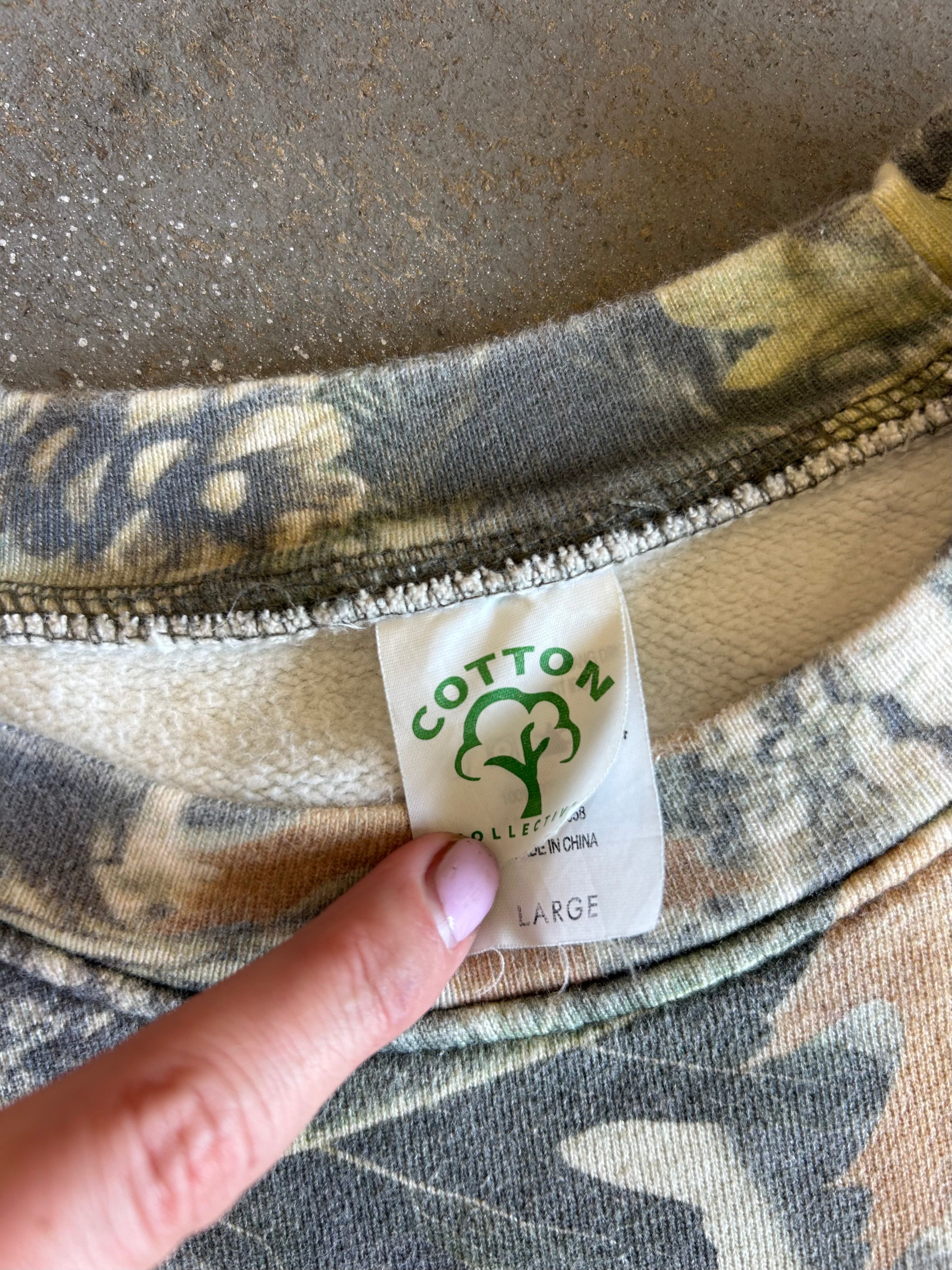 Close-up of a camouflage-patterned garment collar with a visible cotton label reading 'COTTON COLLECTIVE' and a finger pointing at the label.