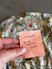 A hand with lavender nail polish holds a peach-colored sticky note with handwritten text reading '- SAMPLE - BACK DESIGN FLAW' in front of a camouflage-patterned garment.