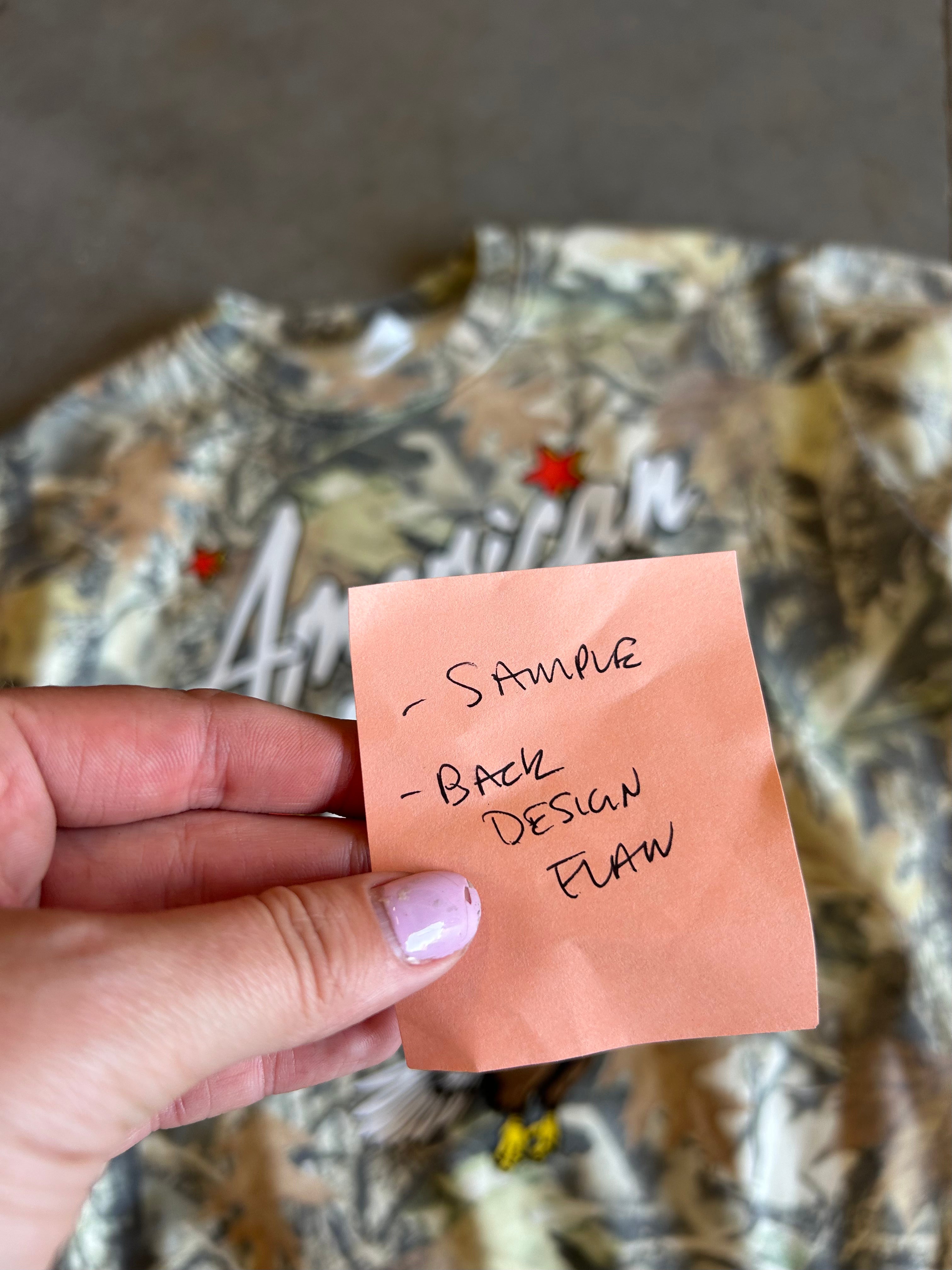 A hand with lavender nail polish holds a peach-colored sticky note with handwritten text reading '- SAMPLE - BACK DESIGN FLAW' in front of a camouflage-patterned garment.