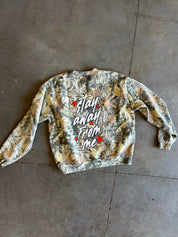 A long-sleeve crewneck sweatshirt with a camouflage pattern in shades of green, brown, and beige, featuring the phrase 'stay away from me' in white script with red star accents on the back. The sweatshirt is laid flat on a gray concrete floor.