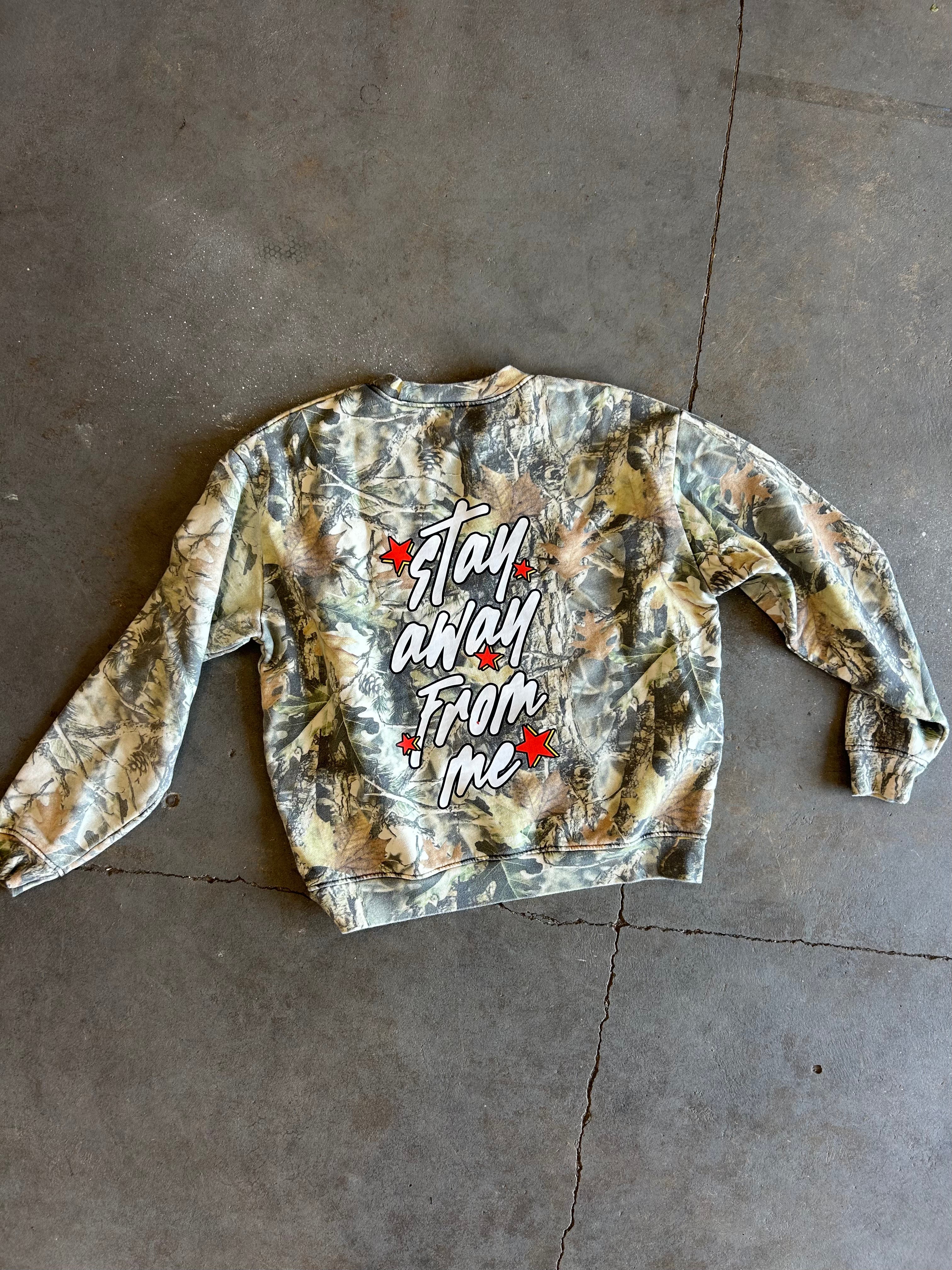 A long-sleeve crewneck sweatshirt with a camouflage pattern in shades of green, brown, and beige, featuring the phrase 'stay away from me' in white script with red star accents on the back. The sweatshirt is laid flat on a gray concrete floor.