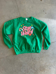 A bright green crewneck sweatshirt with a colorful retro graphic reading 'Stay Weird' in pink and white, decorated with stars, hearts, and a rainbow, laid flat on a concrete floor