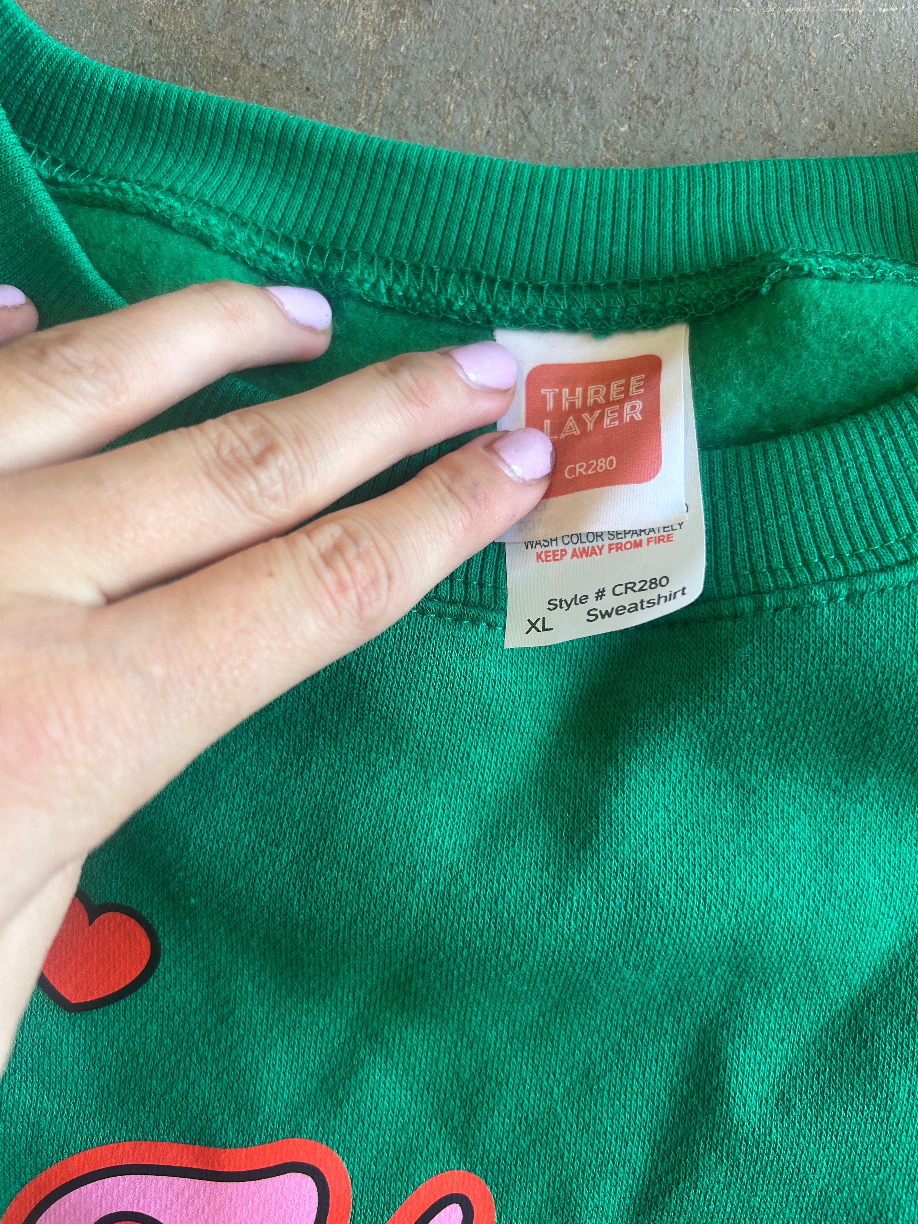 Close-up of a green sweatshirt neck label being held by a hand with light purple nail polish. The label reads THREE LAYER and shows size XL.