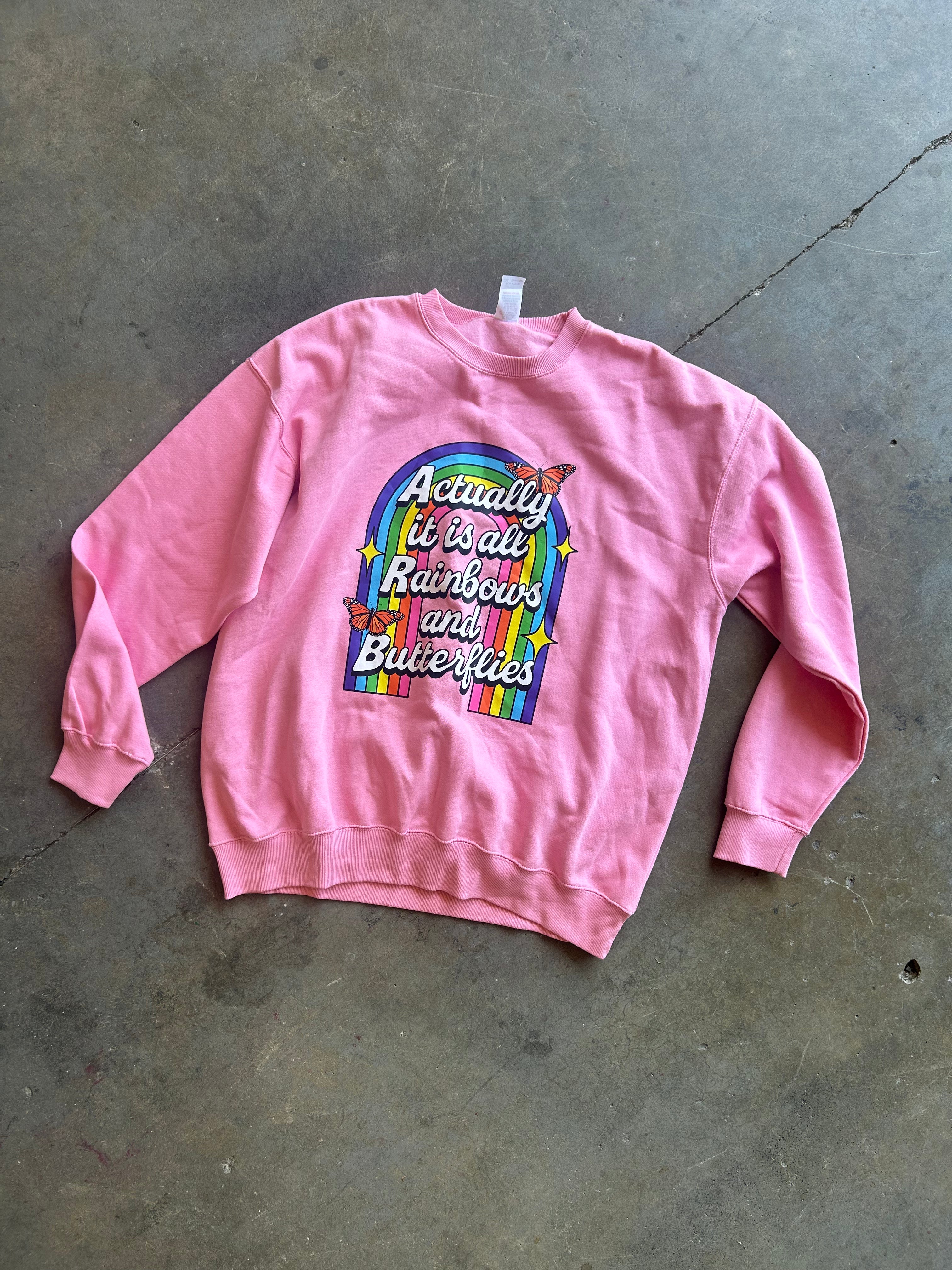 A pink crewneck sweatshirt with a colorful graphic on the front that reads 'Actually it is always Rainbows and Butterflies' in white script over a rainbow arch, laid flat on a gray concrete floor.