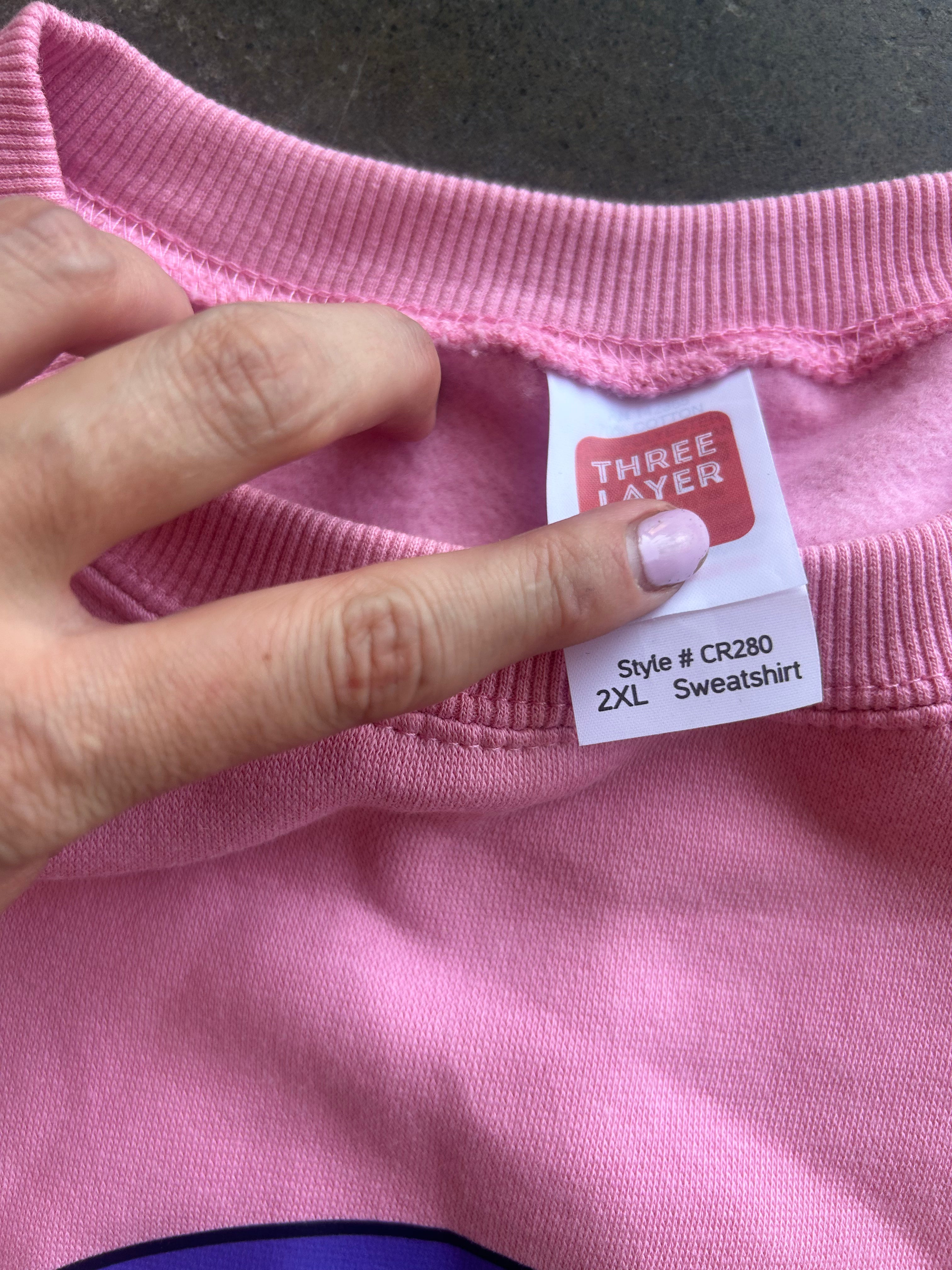 A close-up of a pink sweatshirt showing the inner neck label, which reads 'THREE LAYER' and 'Style # CR280 2XL Sweatshirt'. A hand with light pink nail polish is holding the collar open to display the tag.