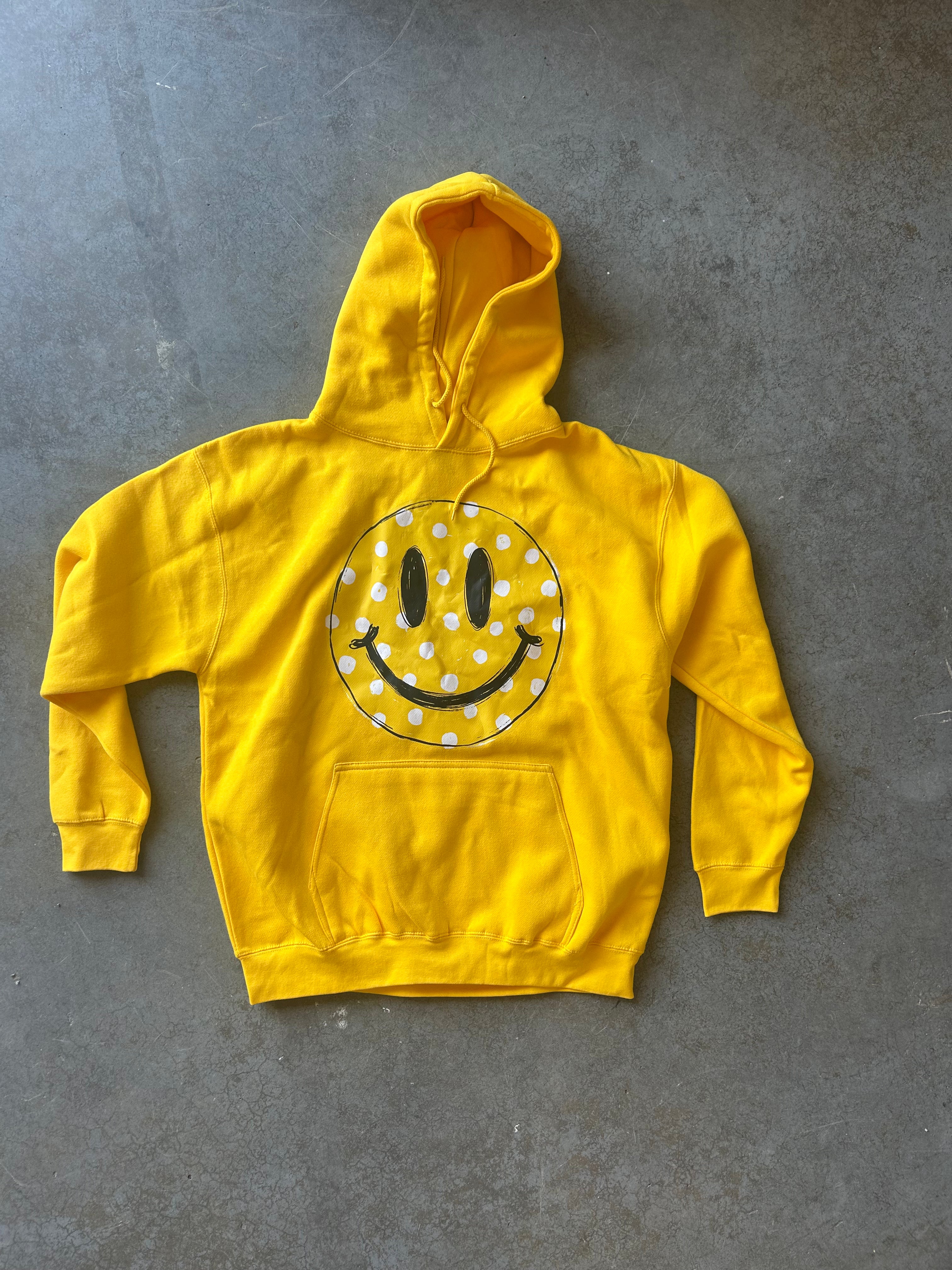 A bright yellow pullover hoodie with a large smiley face graphic on the chest, laid flat on a textured gray concrete surface.