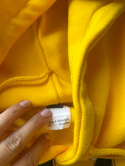 Close-up of a bright yellow hoodie showing the inner neck label, with a hand pointing to it. The label reads 'Size & Midweight Hoodies' and indicates size XL.