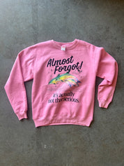 A pink crewneck sweatshirt with a graphic of a jumping fish and the text 'Almost Forgot! it's actually not that serious.' laid flat on a gray concrete floor.
