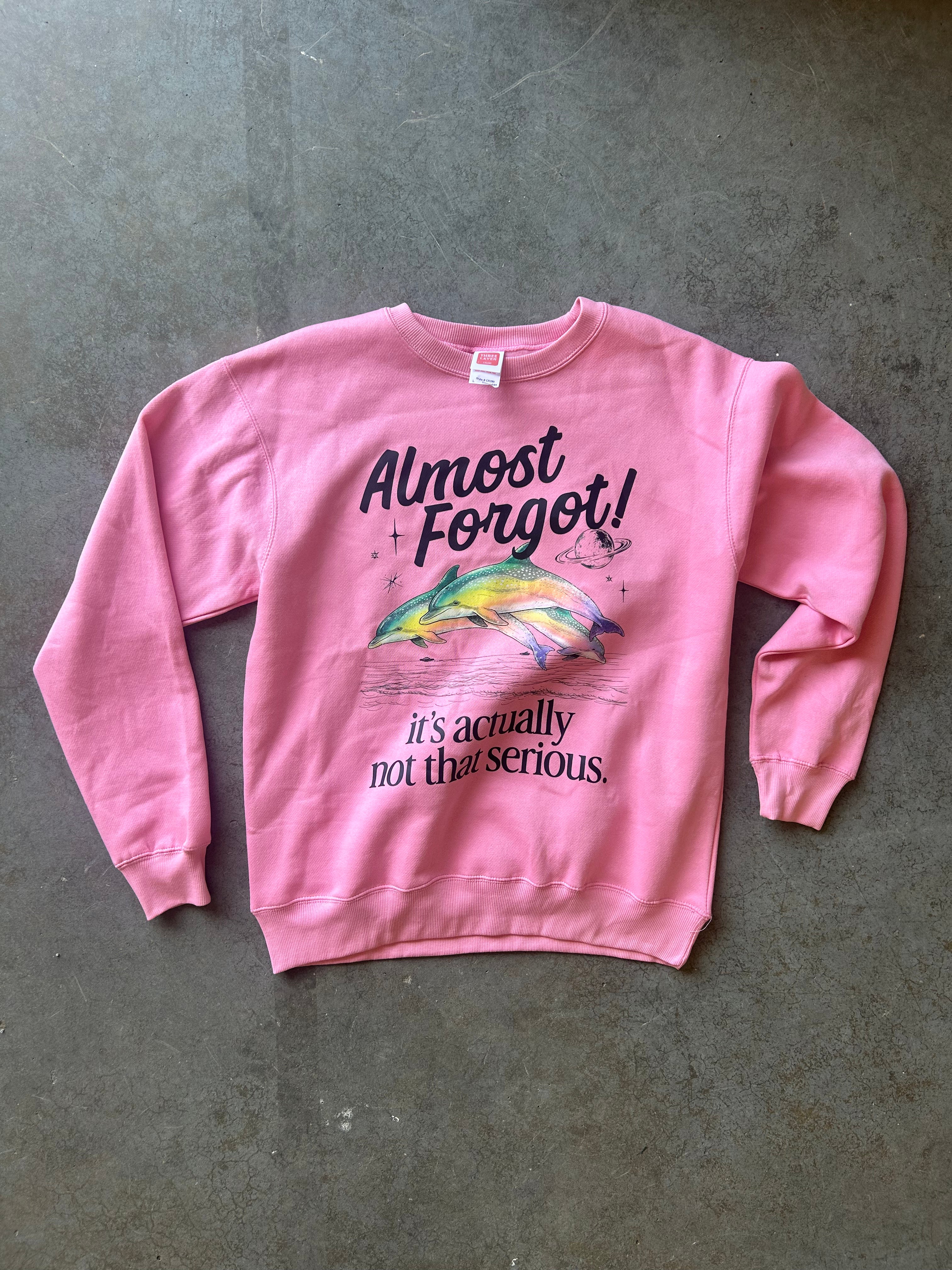 A pink crewneck sweatshirt with a graphic of a jumping fish and the text 'Almost Forgot! it's actually not that serious.' laid flat on a gray concrete floor.
