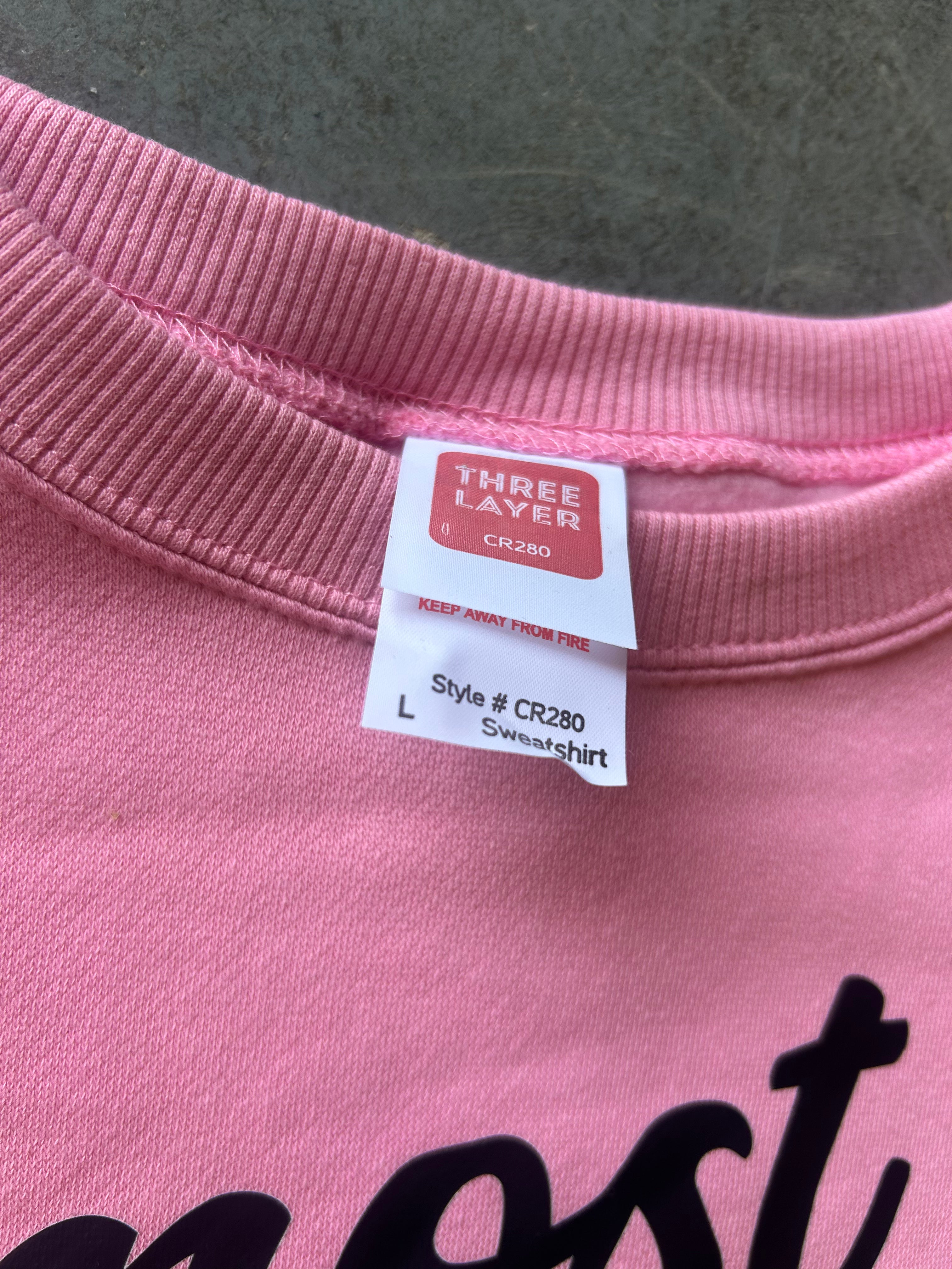Close-up of a pink crewneck sweatshirt showing the inner neck label with 'THREE LAYER' branding and size information, with part of a black graphic print visible at the bottom.