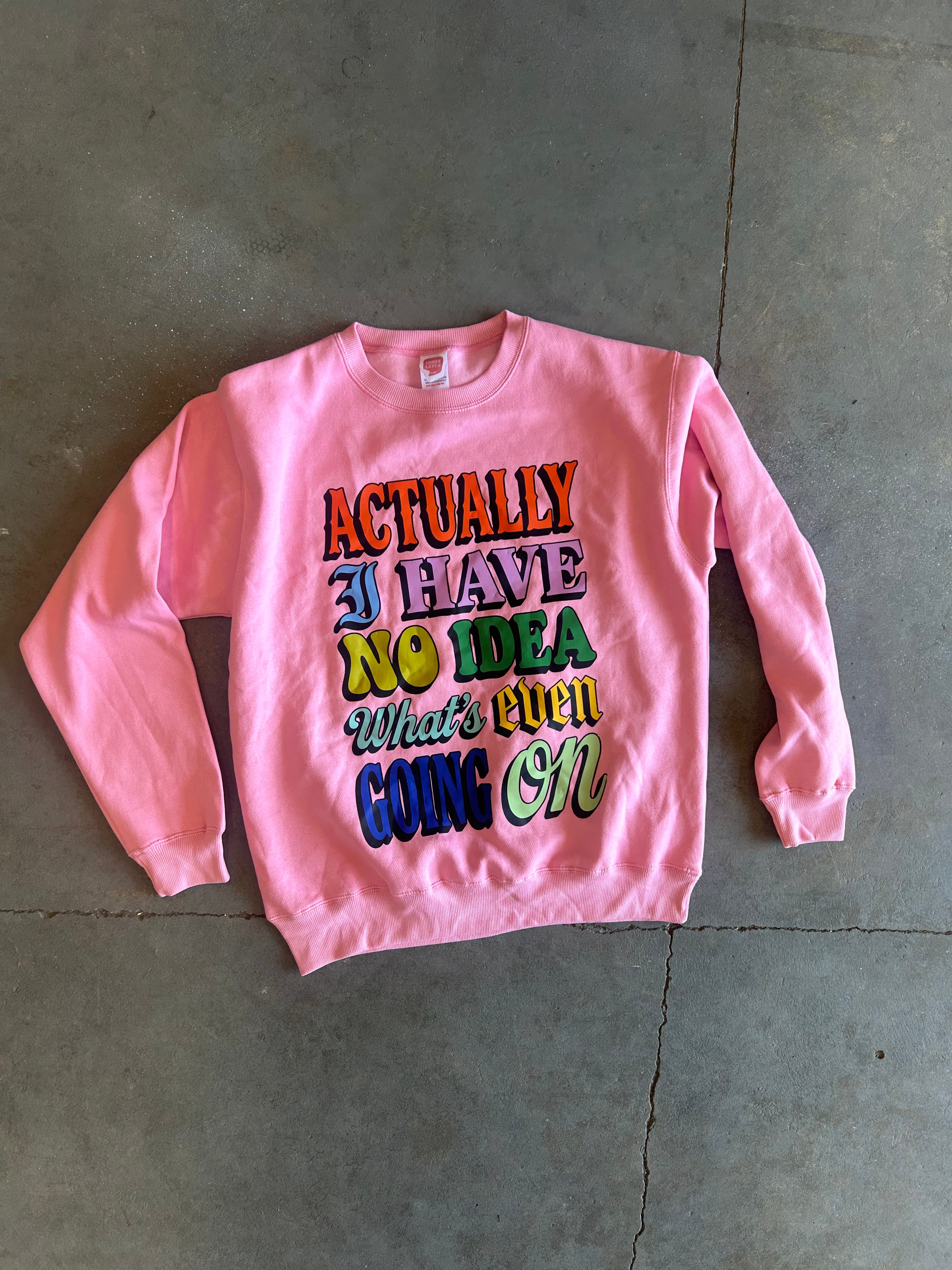 A pink crewneck sweatshirt with a colorful retro-style text graphic reading 'ACTUALLY I HAVE NO IDEA What's even GOING ON', laid flat on a gray concrete floor.