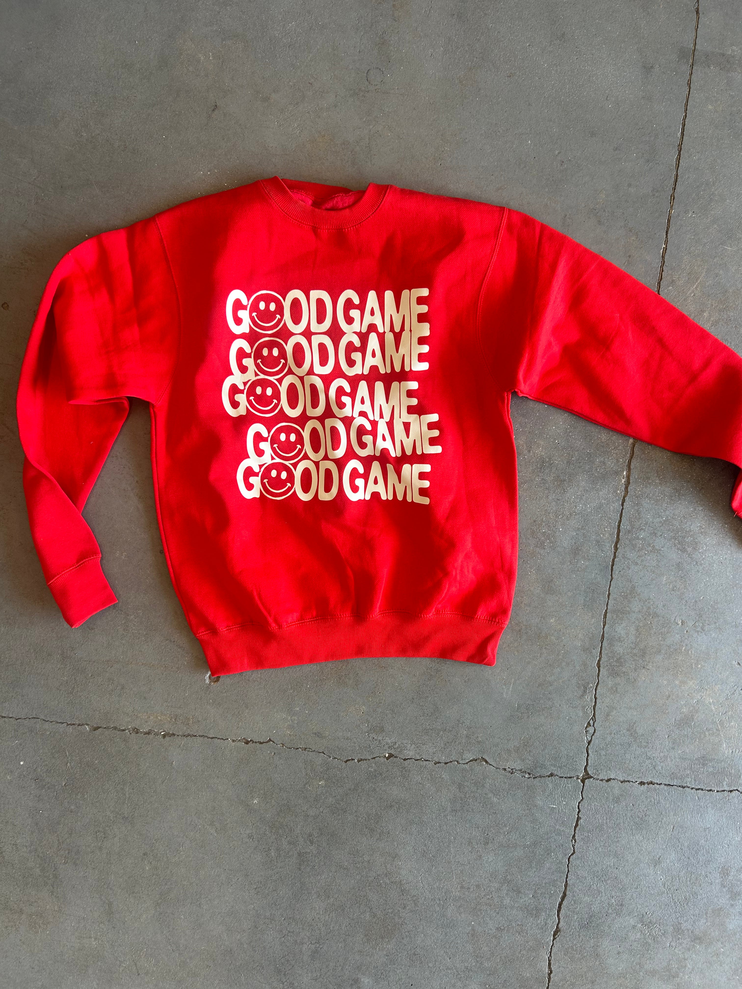 A bright red crewneck sweatshirt with a graphic print of the phrase 'GOOD GAME' repeated vertically in white and gold, featuring a smiley face inside each word. The sweatshirt is laid flat on a gray concrete floor.