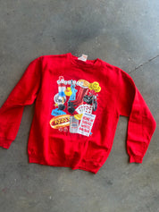 A bright red crewneck sweatshirt with a colorful graphic collage on the front, laid flat on a gray concrete floor.