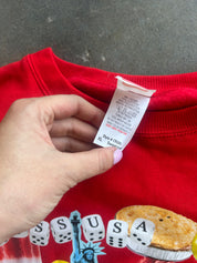 A close-up of a red sweatshirt showing the inner neck label, with a hand holding it up. The sweatshirt has a colorful graphic at the bottom featuring dice spelling 'SSUSA' and a pie.
