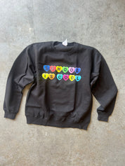 A dark charcoal gray crewneck sweatshirt with the phrase 'SCHOOL IS COOL' printed across the chest in colorful heart-shaped letters, laid flat on a textured concrete floor.