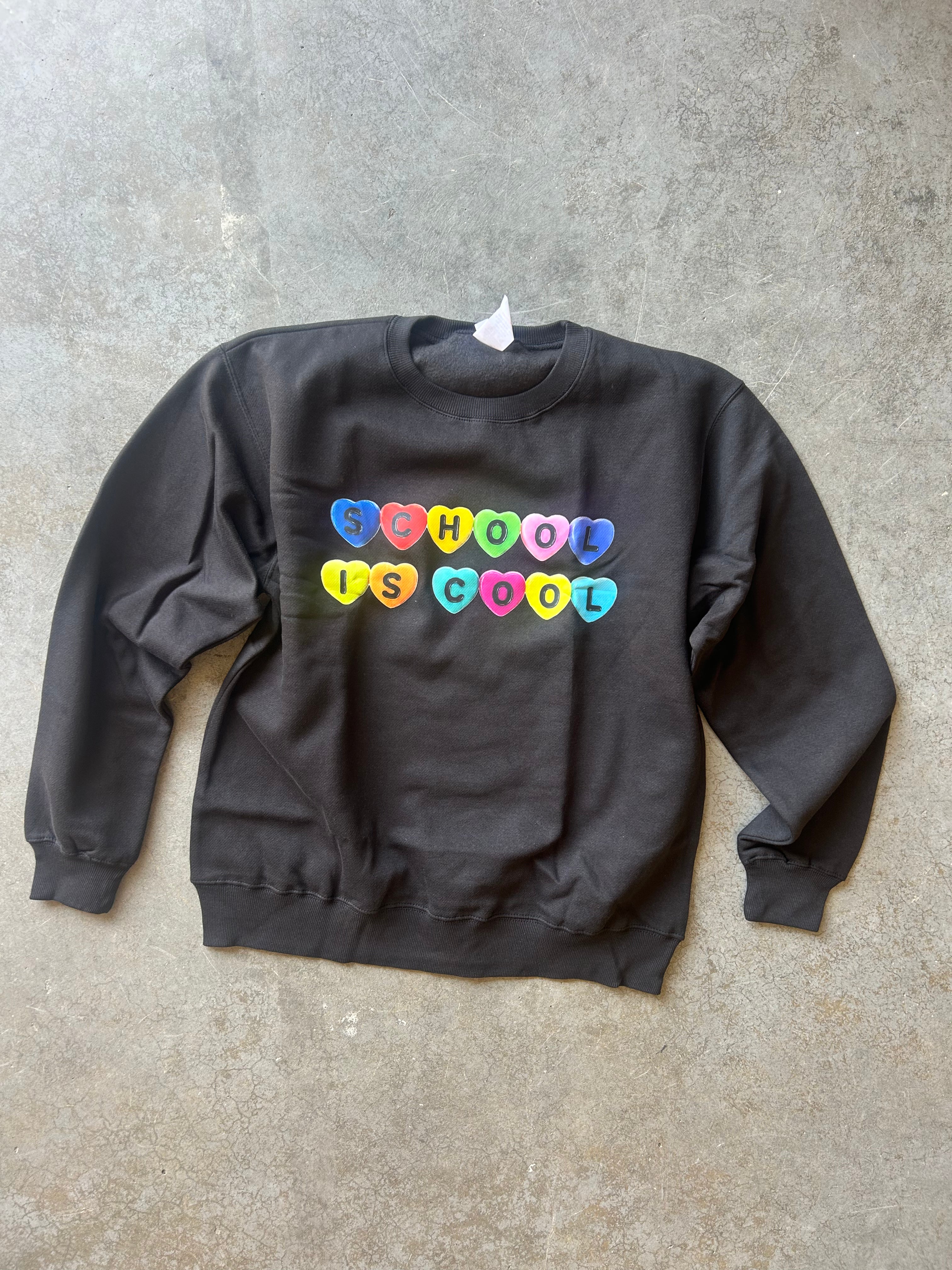 A dark charcoal gray crewneck sweatshirt with the phrase 'SCHOOL IS COOL' printed across the chest in colorful heart-shaped letters, laid flat on a textured concrete floor.