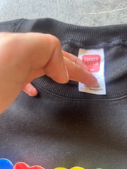 A close-up of a black crewneck sweatshirt showing the inner neck label, which reads 'THREE LAYER'. A hand is pointing at the label.