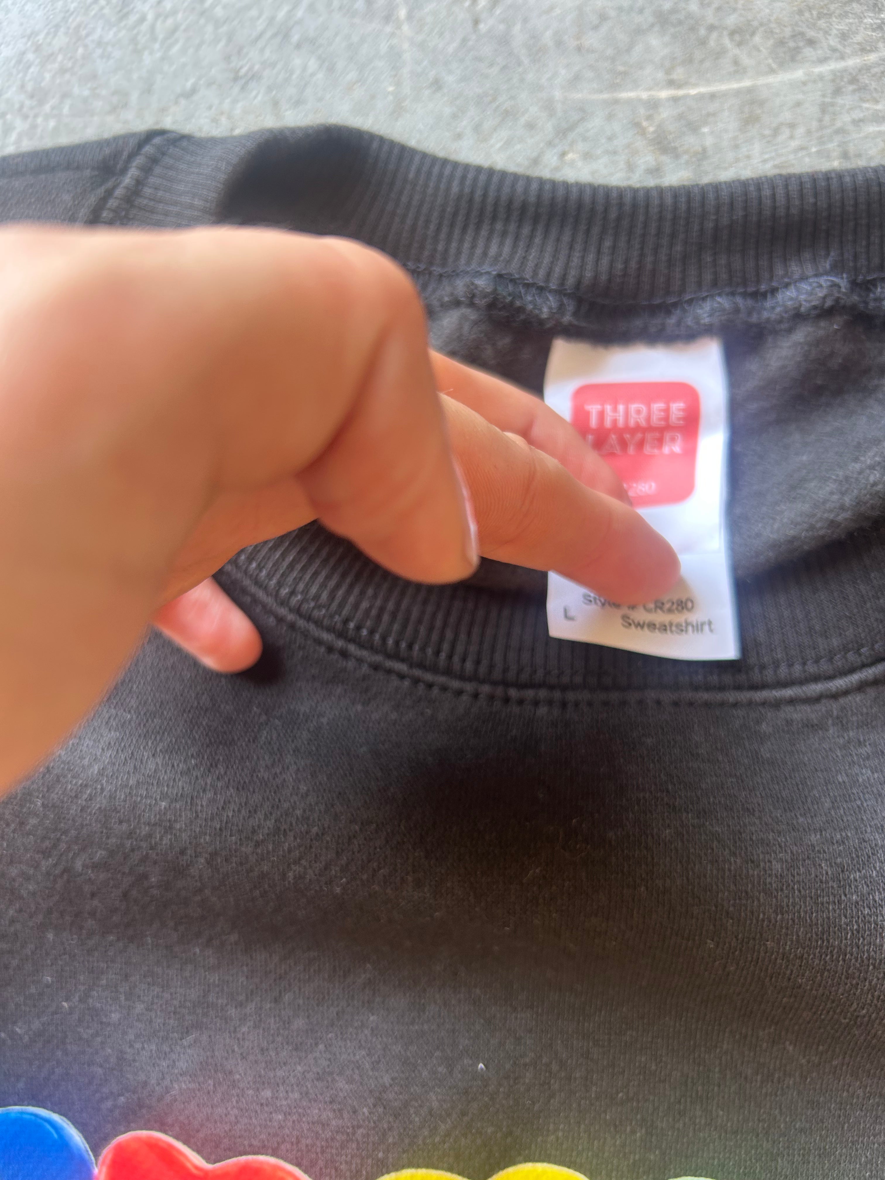 A close-up of a black crewneck sweatshirt showing the inner neck label, which reads 'THREE LAYER'. A hand is pointing at the label.