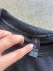 Close-up of a black Gildan cotton blend t-shirt neck tag being held by a hand against a gray concrete background.