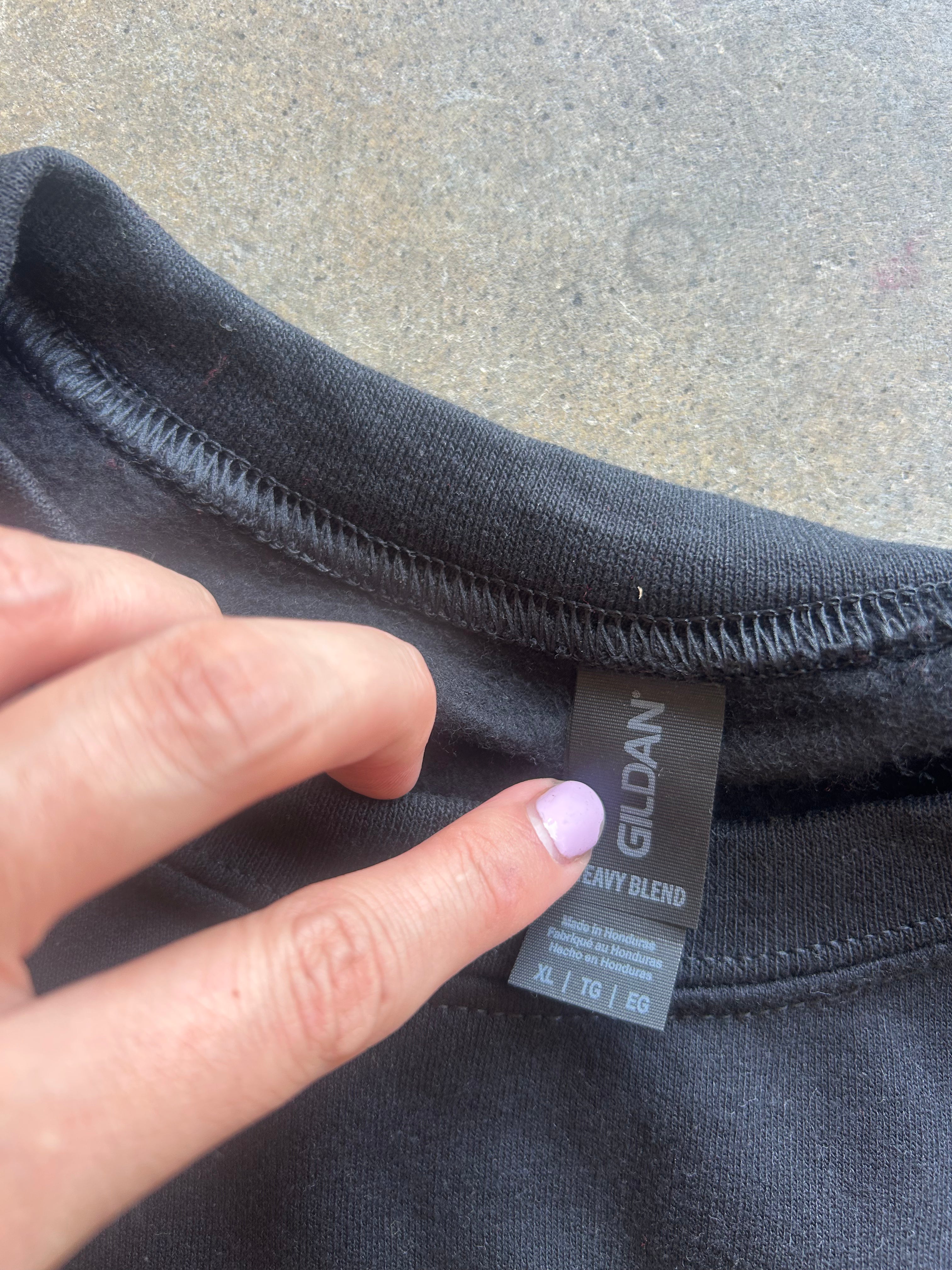 Close-up of a black Gildan cotton blend t-shirt neck tag being held by a hand against a gray concrete background.
