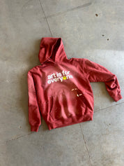 A red hooded sweatshirt with 'art is for everyone' printed on the chest, laid flat on a gray concrete floor.