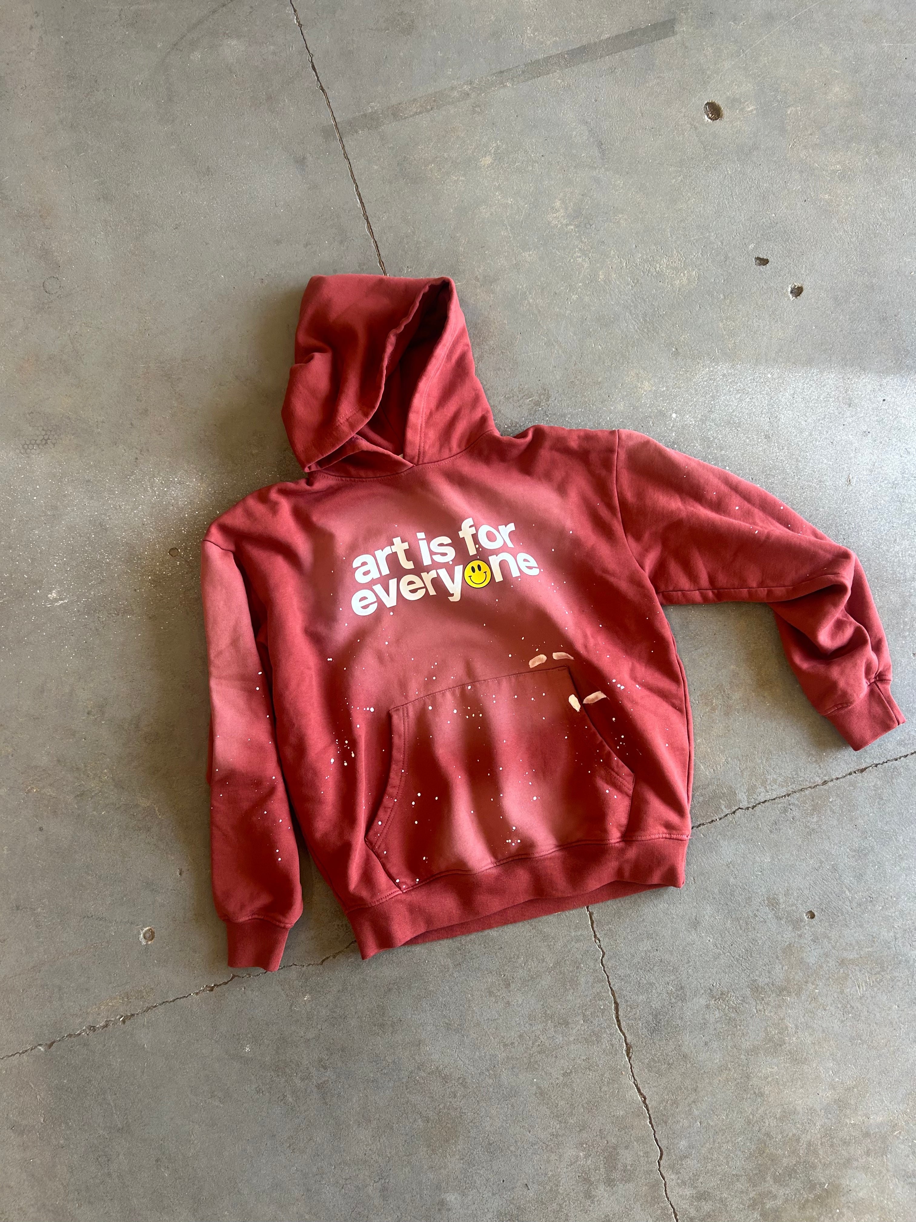 A red hooded sweatshirt with 'art is for everyone' printed on the chest, laid flat on a gray concrete floor.