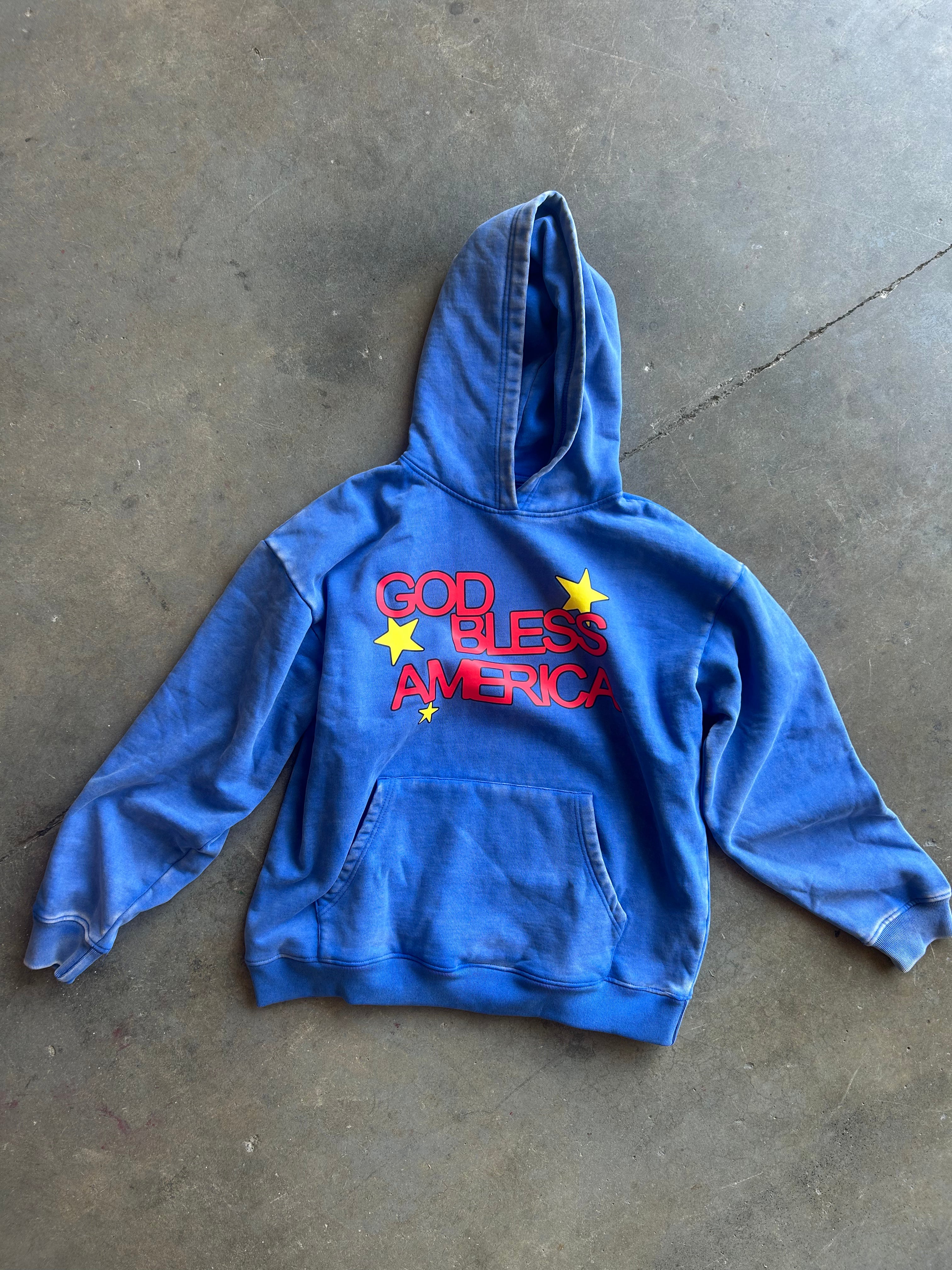 A blue hooded sweatshirt laid flat on a concrete floor, featuring the text 'GOD BLESS AMERICA' in red with yellow stars on the chest.
