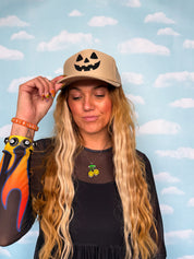 A woman with long blonde wavy hair wearing a beige baseball cap with a black jack-o'-lantern face, posing against a light blue sky and clouds backdrop.