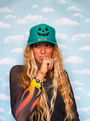 A woman with long blonde wavy hair wearing a green pumpkin jack cap and a black long-sleeve shirt with orange and yellow flame patterns, posing against a light blue sky and clouds backdrop.